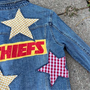 Peut inclure: Veste en jean bleue avec des &eacute;cussons en forme d'&eacute;toile en vichy jaune et vichy rouge et blanc. Le mot "CHIEFS" est imprim&eacute; en rouge sur un fond jaune au dos de la veste.