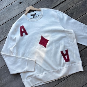 May include: Cream-colored sweatshirt with a crew neck and long sleeves. Features two red "A" appliqués and a red diamond appliqué. The sweatshirt is hanging on a wooden hanger.