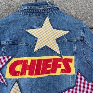 Peut inclure: Veste en jean bleue avec un design patchwork. Le dos de la veste pr&eacute;sente le mot "CHIEFS" en lettres rouges sur un fond jaune, ainsi que plusieurs patchs en forme d'&eacute;toile dans des motifs vichy jaunes et rouges.