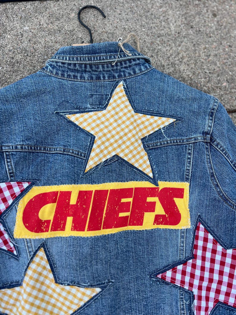 Peut inclure: Veste en jean bleue avec un design patchwork. Le dos de la veste pr&eacute;sente le mot "CHIEFS" en lettres rouges sur un fond jaune, ainsi que plusieurs patchs en forme d'&eacute;toile dans des motifs vichy jaunes et rouges.