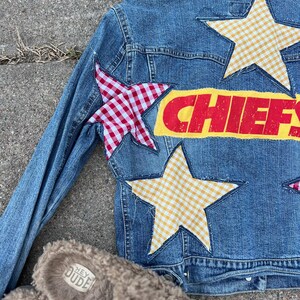 Peut inclure: Veste en jean bleue orn&eacute;e d'&eacute;toiles en vichy rouge et blanc et en vichy jaune. Le mot "CHIEFS" est imprim&eacute; en rouge sur un fond jaune. Une paire de pantoufles marron floues avec le texte "HEY DUDE" est au premier plan.