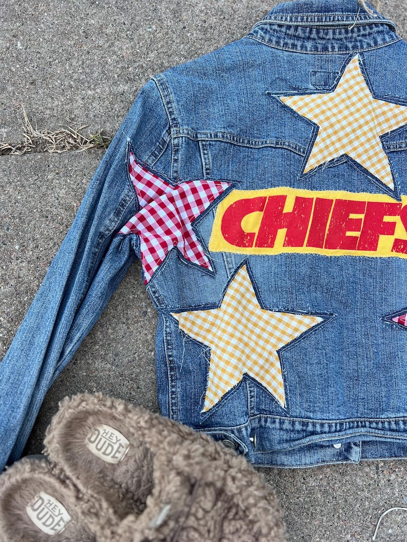 Peut inclure: Veste en jean bleue orn&eacute;e d'&eacute;toiles en vichy rouge et blanc et en vichy jaune. Le mot "CHIEFS" est imprim&eacute; en rouge sur un fond jaune. Une paire de pantoufles marron floues avec le texte "HEY DUDE" est au premier plan.