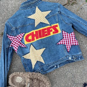 May include: A light blue denim jacket with the word "CHIEFS" in red letters on a yellow background. The jacket is decorated with gingham and yellow star patches. A pair of brown fuzzy slippers with the text "HEY DUDE" are at the bottom.