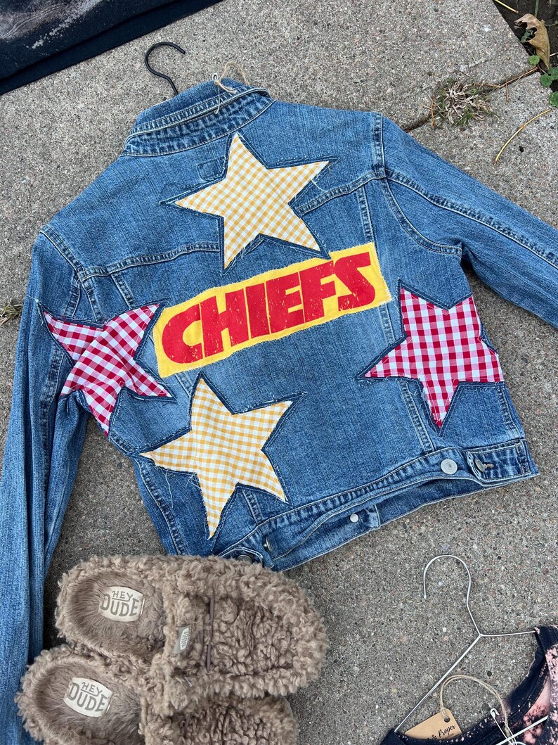 Peut inclure: Veste en jean bleu clair avec le mot "CHIEFS" en lettres rouges sur fond jaune. La veste est orn&eacute;e d'&eacute;toiles &agrave; carreaux et jaunes. Une paire de pantoufles marron et floues avec le texte "HEY DUDE" est en bas.