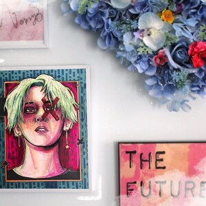 May include: A framed illustration of a person with green hair and a red X over their eye. The background is a vibrant pink and blue with stars. The frame is white with a blue and green pattern. There is a sign below that says "THE FUTURE" in black text on a pink and orange background.