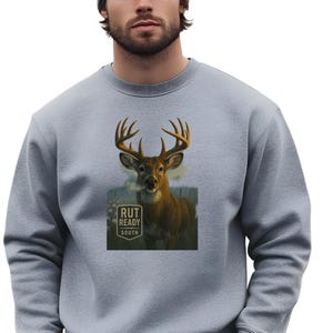 Könnte beinhalten: Ein hellgraues Sweatshirt mit einer detaillierten Illustration eines Hirsches mit großem Geweih. Das Reh ist vor einem Hintergrund platziert, mit dem Text "RUT READY SOUTH" in einem Quadrat. Die Person im Bild trägt eine graue Mütze.