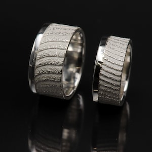 May include: Two silver wedding bands with a textured, wavy design. The bands are both wide and have a polished edge.