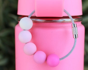 May include: A pink water bottle with a gray cable loop attached to the lid. The loop has five pink and white beads attached to it.