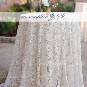 May include: A white lace tablecloth with a delicate floral pattern covers a round table. The tablecloth is draped over the table and hangs down to the floor. The table is set for a wedding or other special event.