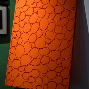 Può includere: Opera d'arte astratta arancione con un motivo di cerchi irregolari rosso scuro. L'opera è esposta su una sedia nera, con uno sfondo di parete verde e bianca. Il pezzo è una tela rettangolare.