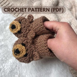Puede incluir: Un par de botines de bebé de crochet marrón con caras de oso. Los botines tienen ojos negros y narices marrones.