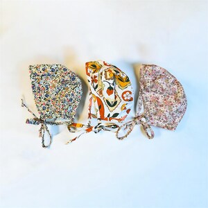 May include: Three baby bonnets in various floral and patterned designs. The bonnet on the left features a blue, pink, and yellow floral print. The middle bonnet has an orange, yellow, and white pattern with strawberries. The right bonnet has a pink floral pattern.