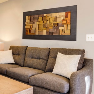 The wooden wall art is on the sofa in a mid-century style living room. Its dimensions are: 48 x 24 in. The interior has an embossed composition of recycled wood pieces of different shapes and tones. Around it is a narrow area painted dark brown.