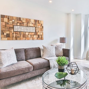 May include: A modern living room with a brown sofa, decorative pillows, and a glass-topped coffee table. A large wooden art piece hangs above the sofa. A white armchair with a geometric pillow sits near a window.