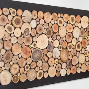 Puede incluir: Un cuadro rectangular compuesto por un mosaico de numerosas rodajas de madera circulares de diversos tama&ntilde;os y tonos marrones naturales, dispuestas dentro de un marco negro s&oacute;lido. Las piezas de madera muestran vetas y anillos visibles.
