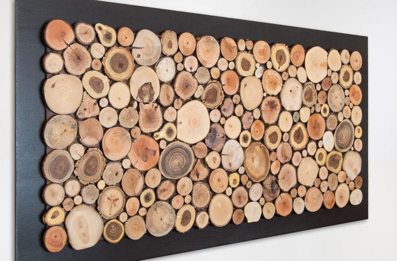 Puede incluir: Un cuadro rectangular compuesto por un mosaico de numerosas rodajas de madera circulares de diversos tama&ntilde;os y tonos marrones naturales, dispuestas dentro de un marco negro s&oacute;lido. Las piezas de madera muestran vetas y anillos visibles.