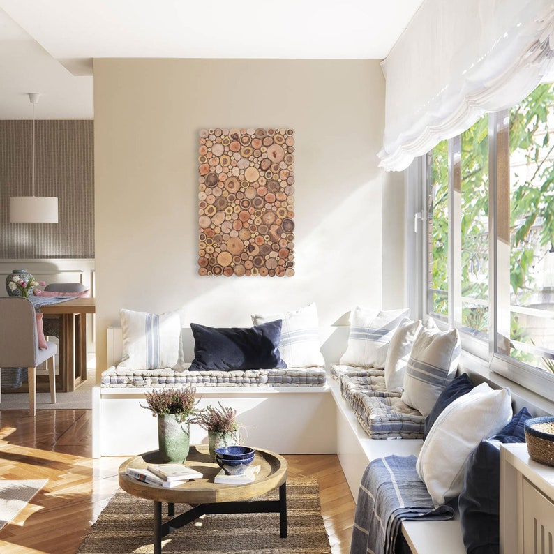 May include: A white couch with blue and white striped pillows sits in front of a wall with a wood slice art piece. The couch is in a living room with a wooden floor and a window with green plants outside.