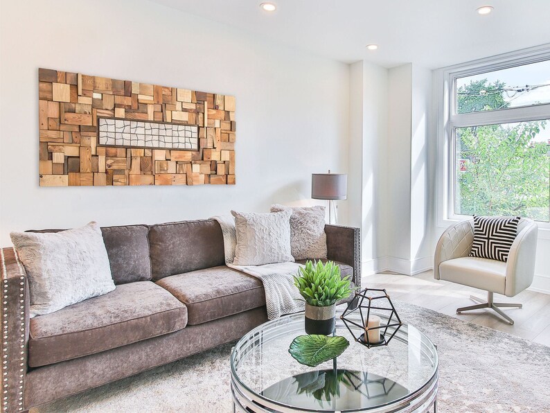May include: A modern living room with a brown sofa, decorative pillows, and a glass-topped coffee table. A large wooden art piece hangs above the sofa. A white armchair with a geometric pillow sits near a window.