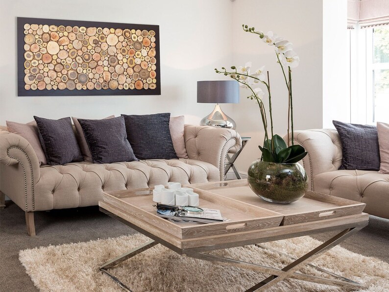 May include: A rectangular coffee table featuring two light wood trays with cutout handles, supported by a silver metal X-frame base. The table holds a glass bowl with a white orchid plant and a small collection of white ceramic objects.