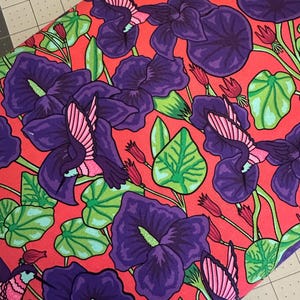 May include: Vibrant fabric featuring a floral pattern with large purple flowers, green leaves, and pink hummingbirds against a bright red background. The design is a repeating pattern, creating a bold and colorful textile.