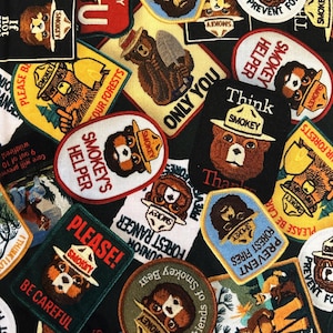 May include: A collection of Smokey Bear patches in various shapes and sizes. The patches feature Smokey Bear in a ranger hat, with text like "Prevent Forest Fires," "Smokey's Helper," and "Please Be Careful." The background is dark green.