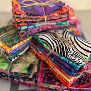 May include: Stacks of colorful fabric squares tied with twine. The fabrics feature a variety of patterns, including floral, zebra stripes, and abstract designs. The color palette includes vibrant hues like pink, orange, purple, green, and blue.