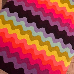 Pode incluir: Um tecido vibrante com um padrão ondulado num espectro de cores. O design apresenta faixas alternadas de rosa, laranja, amarelo, verde, azul, roxo e preto. O tecido é provavelmente usado para projetos de artesanato ou costura.