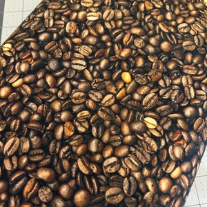 May include: A close-up of fabric patterned with roasted coffee beans. The beans range in colour from light tan to dark brown, with golden yellow highlights. The fabric is folded, and a ruler is visible in the background.