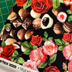 May include: Black fabric with a repeating pattern of red and pink roses and chocolate candies. The fabric has the text "TRUE ROMANCE BY GRETA LYNN" printed on the bottom edge.