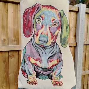 Op de afbeelding: Een beige draagtas met een kleurrijk teckelontwerp. De oren van de hond zijn rood en groen, met blauwe, paarse en gele accenten. De tas heeft een groene rand en een handvat.