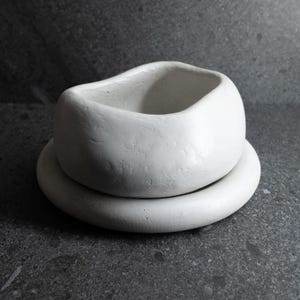 Könnte beinhalten: Eine kleine, handgefertigte, weiße Keramikschale mit abgerundeter Form, die auf einem passenden, flachen, kreisförmigen Sockel steht. Die Schale hat eine glatte, matte Oberfläche und einen leicht unregelmäßigen Rand. Das Design ist minimalistisch und modern.
