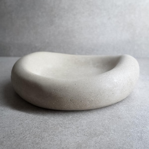 Puede incluir: Un cuenco de hormigón gris claro, redondeado, con interior liso y exterior texturizado. El cuenco tiene una forma ovalada poco profunda y está diseñado para uso decorativo o funcional. El color neutro y el diseño minimalista lo hacen adecuado para varios estilos de decoración.