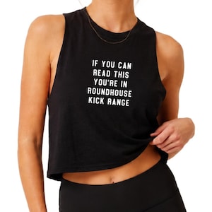 May include: A black sleeveless crop top featuring the white text IF YOU CAN READ THIS YOU'RE IN ROUNDHOUSE KICK RANGE printed on the front. The garment has a crew neckline and wide arm openings, paired with black leggings.