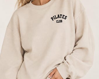 May include: A beige crewneck sweatshirt featuring the text "PILATES CLUB" printed in black block lettering on the upper left chest. The garment has long sleeves with ribbed cuffs and a ribbed neckline, providing a relaxed fit and simple design.