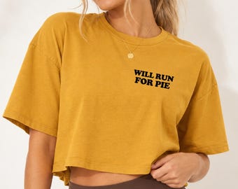 May include: A mustard yellow cropped t-shirt with short sleeves and a crew neckline. The front of the shirt features the black text "WILL RUN FOR PIE" printed in a bold, sans-serif font on the upper left chest area.