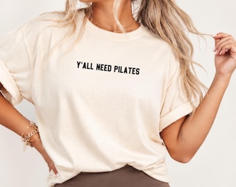 May include: A cream-colored short-sleeve t-shirt with a crew neckline. The front of the shirt features the black text Y'ALL NEED PILATES printed in a simple sans-serif font. The sleeves are rolled up, and the shirt has a relaxed fit.