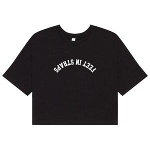 May include: A black cropped t-shirt with short sleeves and a crew neckline. The front of the shirt features the text FEET IN STRAPS printed in a white, arched, collegiate-style font centered across the chest area of the garment.