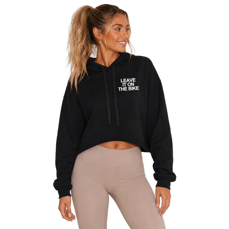 May include: A black cropped hoodie featuring long sleeves, a hood with matching black drawstrings, and the white text "LEAVE IT ON THE BIKE" printed on the upper left chest area. The garment has a relaxed fit and a straight hemline at the waist.