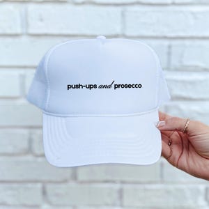 May include: A white trucker hat featuring a foam front panel and a mesh back. The text "push-ups and prosecco" is printed in black on the front panel in a combination of sans-serif and italicized script fonts. The hat has a curved brim.