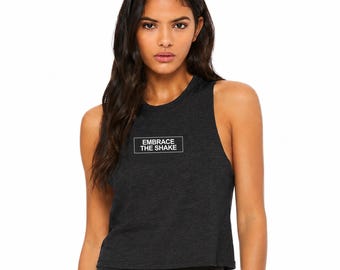 Puede incluir: Un top corto sin mangas de color gris carbón con cuello redondo y un recuadro gráfico blanco rectangular en el pecho que contiene el texto negro EMBRACE THE SHAKE. La prenda está confeccionada con un tejido jaspeado de textura visible.