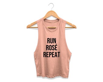 May include: A dusty rose colored sleeveless crop top hanging on a wooden hanger. The shirt features the black text RUN ROSÉ REPEAT printed in a bold, sans-serif font across the center of the chest. The garment has a crew neckline and raw edges.