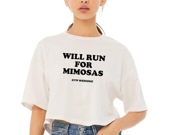 May include: A white short-sleeved cropped t-shirt featuring the black text WILL RUN FOR MIMOSAS in a bold, rounded font, with the smaller text GYM WEEKEND printed underneath, paired with blue denim jeans with a visible metal button and pocket.