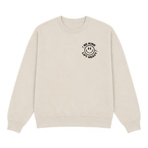 May include: A beige crewneck sweatshirt featuring a black graphic on the upper left chest. The graphic consists of a circular design with a smiley face in the center, surrounded by the text "BE KIND" and "LIFT HEAVY" with small star accents.