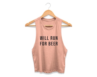 May include: A dusty rose sleeveless crop top with a crew neckline, hanging on a wooden hanger. The text WILL RUN FOR BEER is printed in black, sans-serif capital letters across the center of the chest of the garment.