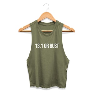 May include: An olive green sleeveless crop top hanging on a wooden hanger. The shirt features the white text 13.1 OR BUST printed across the chest in a clean, sans-serif font. The garment has a round neckline and wide arm openings.