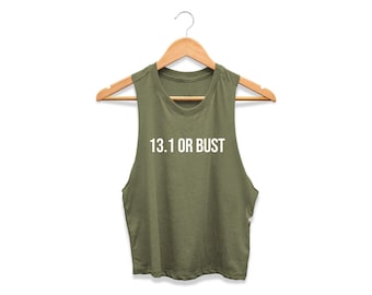 May include: An olive green sleeveless crop top hanging on a wooden hanger. The shirt features the white text 13.1 OR BUST printed across the chest in a clean, sans-serif font. The garment has a round neckline and wide arm openings.