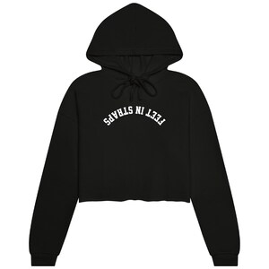 May include: A black cropped hoodie featuring long sleeves, a hood with matching black drawstrings, and the white text FEET IN STRAPS printed in an arched, collegiate-style font across the center of the chest area on the front of the garment.