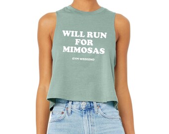 May include: A sage green sleeveless crop top featuring the white text WILL RUN FOR MIMOSAS and the smaller text GYM WEEKEND printed on the front. The garment has a crew neckline and a relaxed fit, paired with light blue denim jeans.