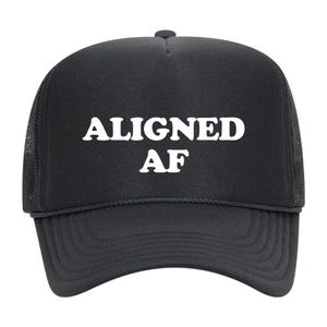 May include: A black trucker hat featuring a foam front panel and mesh back panels. The text ALIGNED AF is printed in bold white capital letters across the center of the front panel. The hat includes a matching black brim with decorative stitching.
