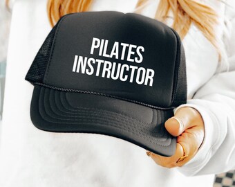 Puede incluir: Una gorra tipo camionero de color negro con panel frontal de espuma y parte trasera de malla. En la parte delantera presenta el texto PILATES INSTRUCTOR en letras mayúsculas blancas y negritas. Incluye una visera negra con costuras decorativas.